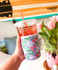 A hand holds a clear plastic cup with a floral-patterned sleeve with paw prints in a bright indoor setting. The cup contains a pink drink and has a pink straw.
