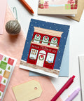 A Christmas-themed greeting card featuring an illustration of a red 'Milk 'n' Cookie Co.' storefront under a snowy blue sky, styled in a flat lay with pastel paper, pens, a glitter ornament, and gift tags.