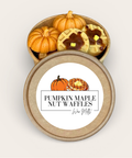 A round kraft paper gift box shown open, with miniature pumpkin-shaped items and nut-waffle shaped wax melts inside. The lid is placed in front and features a white label with the text 'PUMPKIN MAPLE NUT WAFFLES'.