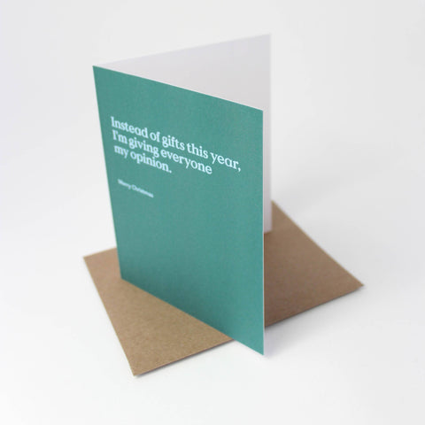 A teal greeting card with white text reading 'Instead of gifts this year, I'm giving everyone my opinion.' and 'Merry Christmas,' shown standing upright with a brown kraft envelope behind it on a white background.