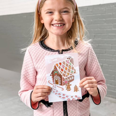 girl holding gingerbread house holiday card with candy looking stickers for decorating the gingerbread house