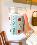 Hand holding a clear cup with a colorful sleeve featuring apples and patterns, against a blurred indoor background. Showing an iced coffee koozie for fall.