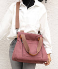 A person wearing a white button-up shirt and gray jeans carries a dusty rose canvas tote bag with tan straps and trim against a textured off-white wall.