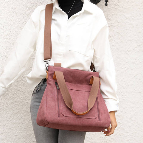 A person wearing a white button-up shirt and gray jeans carries a dusty rose canvas tote bag with tan straps and trim against a textured off-white wall.