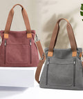 Two canvas tote bags with brown shoulder straps are displayed on a white platform and surface against a light beige wall. The left bag is dusty rose pink and the right bag is gray, both with brown leather-like corner patches and front zippered pockets.