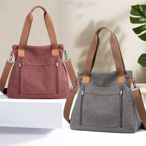 Two canvas tote bags with brown shoulder straps are displayed on a white platform and surface against a light beige wall. The left bag is dusty rose pink and the right bag is gray, both with brown leather-like corner patches and front zippered pockets.