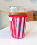 A striped fabric coffee cup sleeve in shades of red, pink, green and white, holding a plastic-wrapped iced coffee on a white tabletop with a softly blurred indoor background.