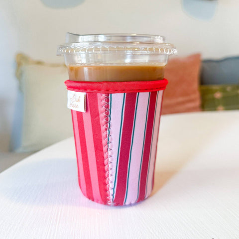 A striped fabric coffee cup sleeve in shades of red, pink, green and white, holding a plastic-wrapped iced coffee on a white tabletop with a softly blurred indoor background.