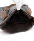 An open gray and brown canvas bag with a white-gloved hand lifting the top flap to show the interior compartments, photographed on a white background.