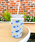 Blue iced beverage sleeve with lemon and blueberry pattern on a wooden table with a brick wall and plants in the background