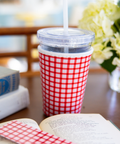 Red and white checkered coffee sleeve around a cup with straw on a table next to an open book and flowers.