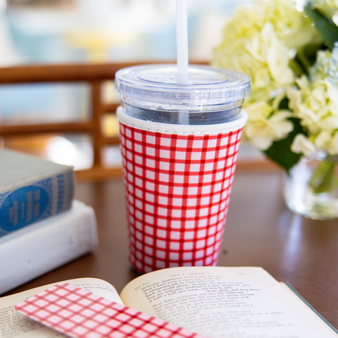 Red and white checkered coffee sleeve around a cup with straw on a table next to an open book and flowers.