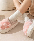 White slippers with pink fuzzy sole worn by a person sitting on a beige surface.