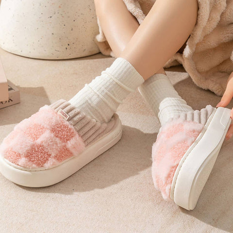 White slippers with pink fuzzy sole worn by a person sitting on a beige surface.