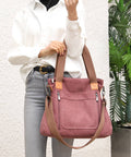 A person wearing a white button-up shirt and light gray jeans holds a dusty rose canvas tote bag with tan straps and trim against a white textured wall next to a potted plant.