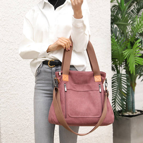 A person wearing a white button-up shirt and light gray jeans holds a dusty rose canvas tote bag with tan straps and trim against a white textured wall next to a potted plant.