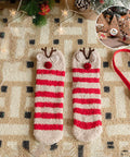 A pair of cozy knit socks with red and cream horizontal stripes and brown reindeer antlers with red noses, displayed on a patterned knit blanket with Christmas decorations and a small circular inset showing a Christmas tree ornament.