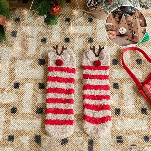 A pair of cozy knit socks with red and cream horizontal stripes and brown reindeer antlers with red noses, displayed on a patterned knit blanket with Christmas decorations and a small circular inset showing a Christmas tree ornament.