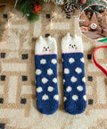 A pair of cozy knit socks with a cat-face design at the top, featuring a navy blue body with white polka dots and a white cat-face cuff. The socks are displayed on a patterned beige rug with Christmas decorations, including a pine cone, evergreen branches, fairy lights, and a small circular inset showing wooden Christmas tree ornaments.