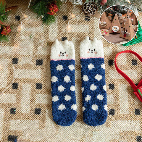 A pair of cozy knit socks with a cat-face design at the top, featuring a navy blue body with white polka dots and a white cat-face cuff. The socks are displayed on a patterned beige rug with Christmas decorations, including a pine cone, evergreen branches, fairy lights, and a small circular inset showing wooden Christmas tree ornaments.