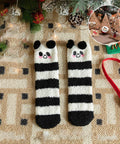 A pair of fuzzy knit socks with black and white horizontal stripes and cute panda faces at the top, laid flat on a beige patterned blanket with Christmas decorations and a small inset photo showing a decorated tree scene.