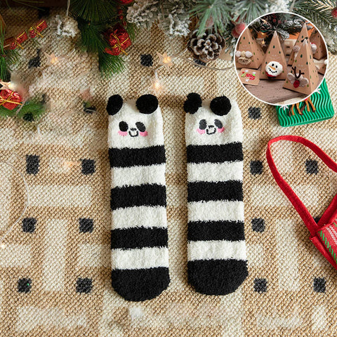 A pair of fuzzy knit socks with black and white horizontal stripes and cute panda faces at the top, laid flat on a beige patterned blanket with Christmas decorations and a small inset photo showing a decorated tree scene.