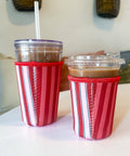 Two insulated travel tumbler cups with clear lids and straws, wrapped in red and white striped sleeve cozies, sitting on a white tabletop.