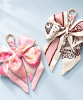 Two bow-shaped silk scarf keychains with gold-tone rings, displayed on a light blue background. One keychain features a pink and cream watercolor pattern, and the other features a cream base with brown graphic patterns and pink stripes.