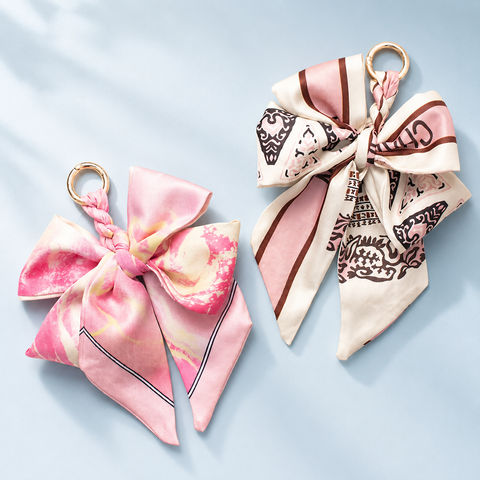 Two bow-shaped silk scarf keychains with gold-tone rings, displayed on a light blue background. One keychain features a pink and cream watercolor pattern, and the other features a cream base with brown graphic patterns and pink stripes.