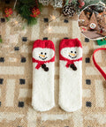 A pair of white knit socks with red cuff trim and a snowman face design, laid flat on a beige patterned blanket with holiday decorations around them. A circular inset shows the socks worn on small wooden trees.