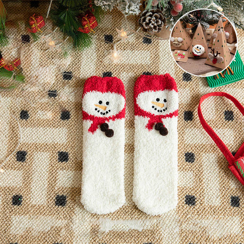 A pair of white knit socks with red cuff trim and a snowman face design, laid flat on a beige patterned blanket with holiday decorations around them. A circular inset shows the socks worn on small wooden trees.