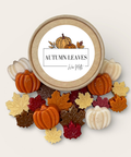 assorted fall wax melts in various shapes like leaves and pumpkins sprinkled on a white background surrounding their kraft colored container