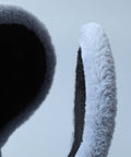 Close-up of a plush faux-fur ear warmer with a dark charcoal interior and a light gray outer edge, shown against a pale blue-gray background.