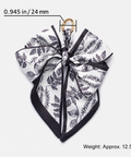 A square scarf tied into a large bow, featuring a black botanical leaf pattern on a white background with black borders. A small gold ring is attached at the top.