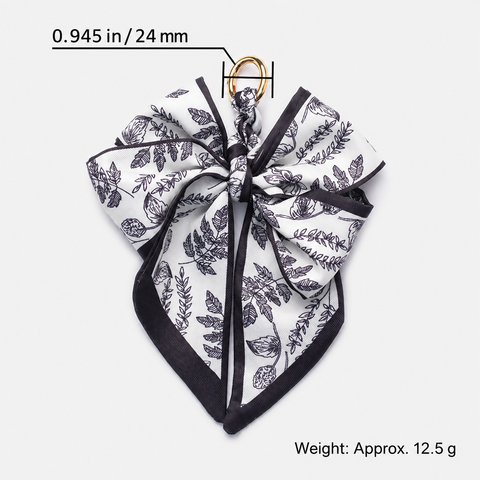 A square scarf tied into a large bow, featuring a black botanical leaf pattern on a white background with black borders. A small gold ring is attached at the top.