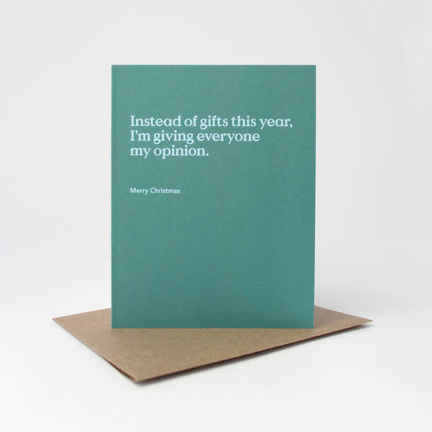 A teal greeting card with white text reading, 'Instead of gifts this year, I'm giving everyone my opinion.' and 'Merry Christmas,' standing upright on a brown kraft envelope against a light gray background.