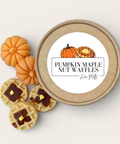 A round kraft paper tin labeled 'Pumpkin Maple Nut Waffles' by Win Motif, shown with three waffle-shaped wax melts topped with maple syrup and butter, alongside three small orange pumpkins on a light beige background.