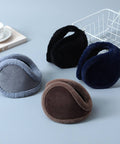 Four pairs of plush earmuffs in different colors—gray, black, navy blue, and brown—are arranged on a light blue-gray surface with styled props, including a white wire basket, a book, and a cup and saucer.